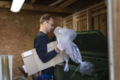 Side view of male blue-collar worker throwing plastic and cardboard waste in garbage bin