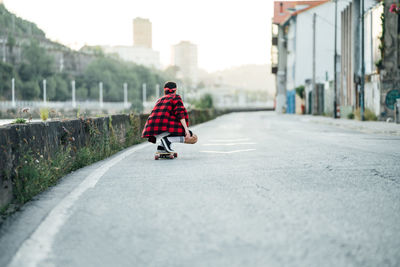 Back view of male hipster in checkered shirt riding longboad along asphalt road while entertaining during weekend in city