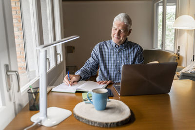 Senior man sitting at home in front of laptop making notes in notebook