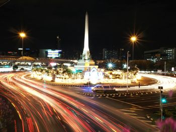 Light trails on road by buildings against sky at night