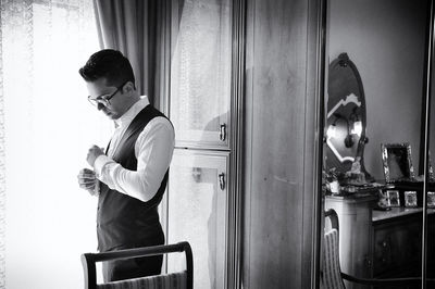 Bridegroom getting dressed while standing by window at home