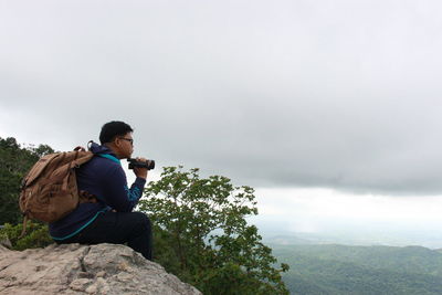 Side view of man with video camera sitting on cliff against cloudy sky
