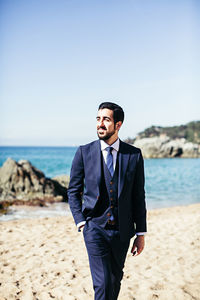 Smiling bridegroom looking away while walking at beach against clear sky during sunny day