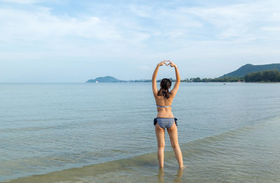 Girl making heart shape at beach against sky