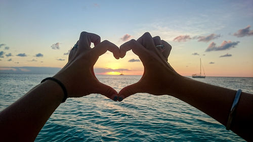 Cropped hands making heart shape by sea against sky during sunset