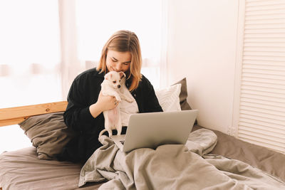 A cheerful young teenage woman plays with her pet a small dog and works using a laptop at home