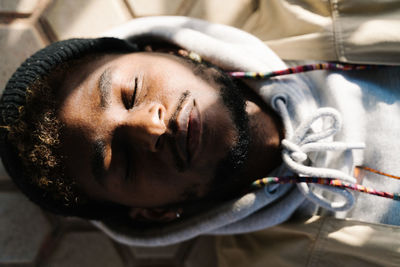 Top view of calm young bearded african american male in trendy outfit lying on ground with closed eyes on sunny day