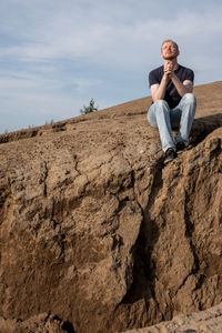 Young man sitting on rock against sky