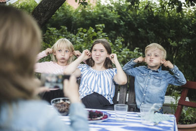 Woman photographing children while making faces at dining table in garden party