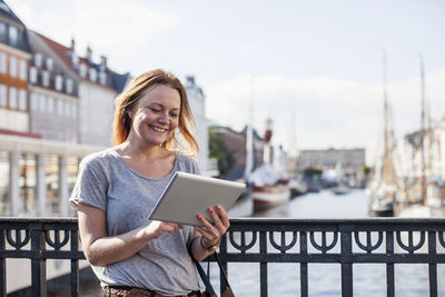 Happy woman using digital tablet against railing at harbor