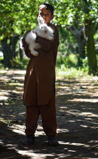 Portrait of young man holding rabbit while standing on road against trees