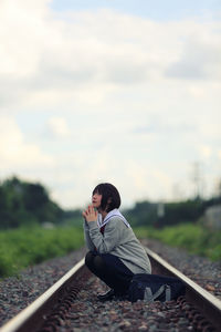 Young woman crouching on railroad track against sky