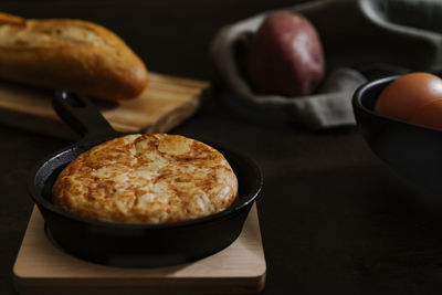 Dark still life picture of a typical spanish potato omelette in a pan with some bread and eggs in the background.