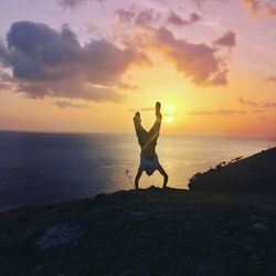 Man doing handstand at beach against sky during sunset