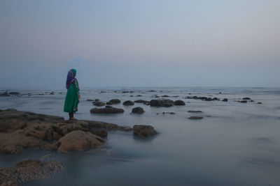 Rear view of woman standing on rock by sea against clear sky