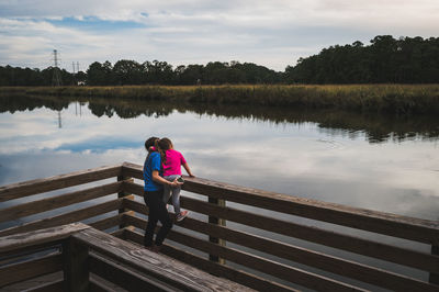 Rear view of woman and daughter standing on wood dock by lake against sky.