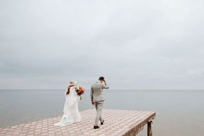 Rear view of newlywed couple walking on pier over lake against cloudy sky