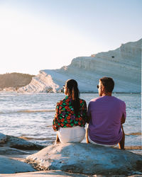 Rear view of women sitting on rock by sea against clear sky