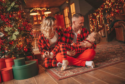 Smile active parents with small son in red checkered sleepwears waiting santa indoor.