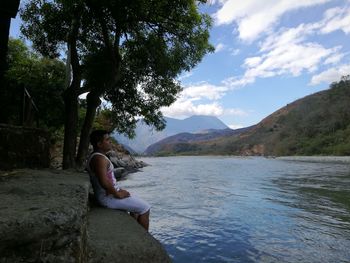 Woman sitting by tree trunk by lake against sky