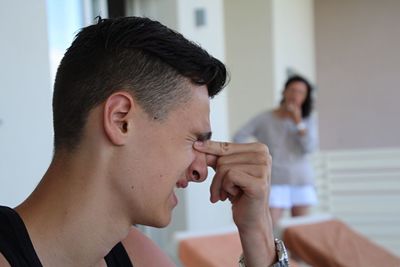 Young man rubbing eyes while mother standing by wall at home