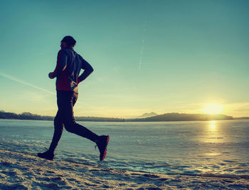 Athlete runner running on beach at frozen lake in rays of sunset. slim man keeps great condition