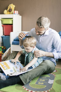 Boy reading book while father combing his hair on floor at home