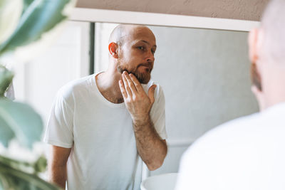 Handsome bald man looking at mirror and touching face in bathroom