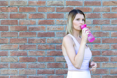 Young woman drinking water while standing against brick wall