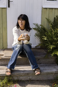 Happy woman holding cup looking away while sitting on porch outside house