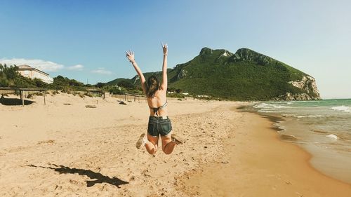 Rear view of woman with arms raised jumping at beach against sky