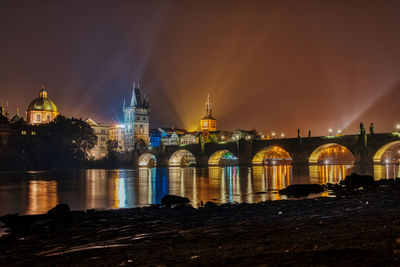 Illuminated bridge over river by buildings against sky at night