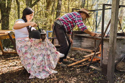 Smiling argentinian female pouring hot water from kettle into calabash gourd against male partner grilling meat above fire in campsite