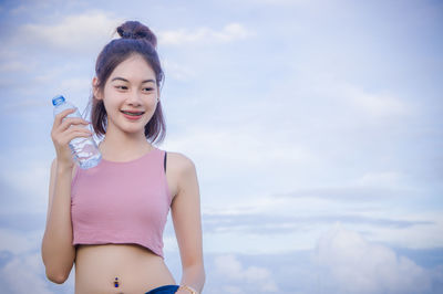 Portrait of smiling young woman with water bottle standing against cloudy sky