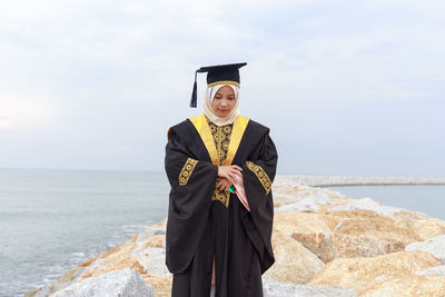 Young woman in graduation gown standing at beach against cloudy sky