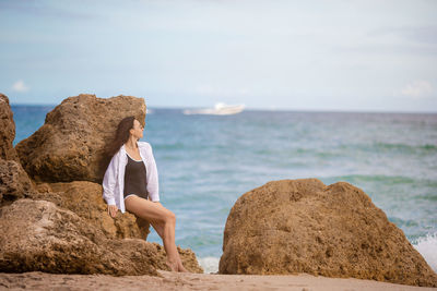 Rear view of woman sitting on rock at beach against sky
