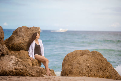 Rear view of woman sitting on rock at beach against sky