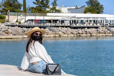 Young woman sitting on shore of sea, wearing black mask. tourism during covid epidemic.