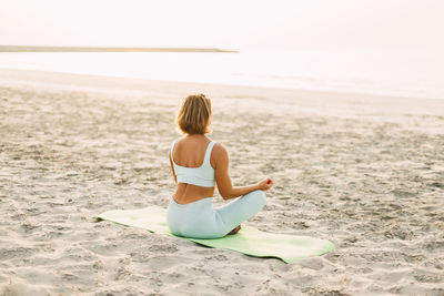 Young woman meditating in pose of lotus on beach near the sea at sunset in summer, rear view