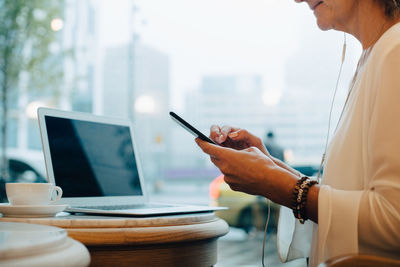 Midsection of businesswoman with laptop on table listening music while sitting by window in cafe