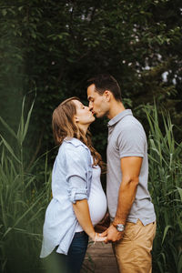 Side view of man kissing pregnant wife while standing amidst plants