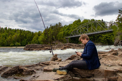 Rear view of man  sitting on rock by the river fishing 