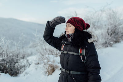 Smiling woman in warm clothing shielding eyes while standing on snowcapped mountain