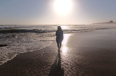 Rear view of woman walking at beach against sky during sunset