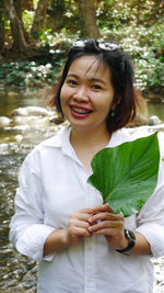 Portrait of cheerful woman holding leaf while standing at riverbank in forest