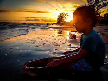 Boy sitting on shore at beach against sky during sunset
