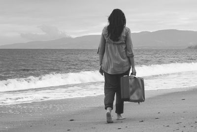 Rear view of woman holding suitcase while walking at beach