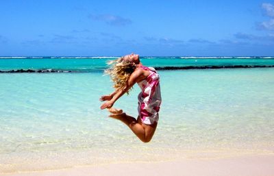 Side view of carefree young woman jumping at beach against sea and sky