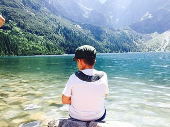 Rear view of boy sitting by lake against mountains