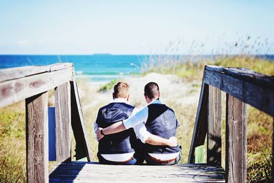 Rear view of men sitting on railing by sea against sky